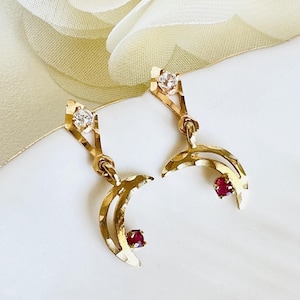 May include: A pair of gold-toned earrings with a crescent moon design. Each earring features a clear, round gemstone at the top and a small, red gemstone at the bottom of the crescent. The earrings are displayed on a white surface.