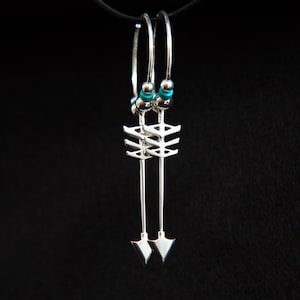 May include: Silver arrow earrings with turquoise beads. These dangle earrings feature a unique design with a geometric arrow shape. The earrings are suspended from silver hoops and are set against a black background.