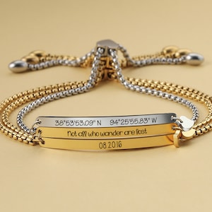 Puede incluir: Un brazalete de oro y plata con cierre deslizante. El brazalete tiene tres barras grabadas con el texto "38°53'53.09" N 94°25′55.83" W", "Not all who wander are lost", y "08.20.16".