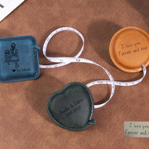 Può includere: Tre metri a nastro retrattili rivestiti in pelle con nastri bianchi. Uno quadrato blu con un'illustrazione di madre e figlio e il testo "To Mom", uno rotondo beige con il testo "I love you, Forever and ever" e uno a forma di cuore verde scuro con "Amélie & Cédric 1996-2026".