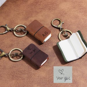 Op de afbeelding: Drie miniatuur notitieboek-sleutelhangers met een leren kaft en antiek messingkleurige hardware. Eén bruin notitieboek is gesloten met 'A.G' gegraveerd, één donkerbruin notitieboek is gesloten met een hart en 'your girl' gegraveerd, en één groen is open.