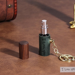 Może przedstawiać: Przenośny atomizer do perfum w ciemnozielonej skórzanej oprawie z wygrawerowanym napisem I Love You z boku. Posiada srebrną końcówkę rozpylającą, odpinany brelok w kolorze złota oraz oddzielną cylindryczną nasadkę z ciemnobrązowego drewna.