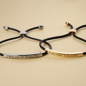 May include: Two adjustable bracelets with black cords and metal bars. One silver bar reads "I Will Love You Always, Dina." The other gold bar displays coordinates. Both bracelets have a sliding knot closure.