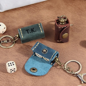 Könnte beinhalten: Drei Würfeltaschen aus Leder mit Metallschlüsselringen und Druckknöpfen. Eine dunkelgrüne Tasche ist mit den Initialen T.K. geprägt, eine ist dunkelbraun und eine ist blau, offen mit zwei weißen Würfeln darin. Drei lose weiße Würfel liegen daneben.