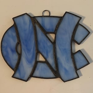 Peut inclure: Une d&eacute;coration de fen&ecirc;tre en vitrail repr&eacute;sentant les lettres "NC" dans un design marbr&eacute; bleu et blanc.