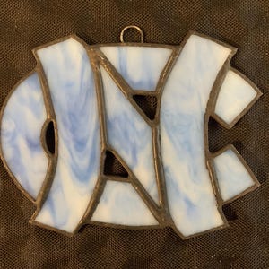 Peut inclure: Une d&eacute;coration en verre teint&eacute; repr&eacute;sentant les lettres "UNC" en bleu et blanc.