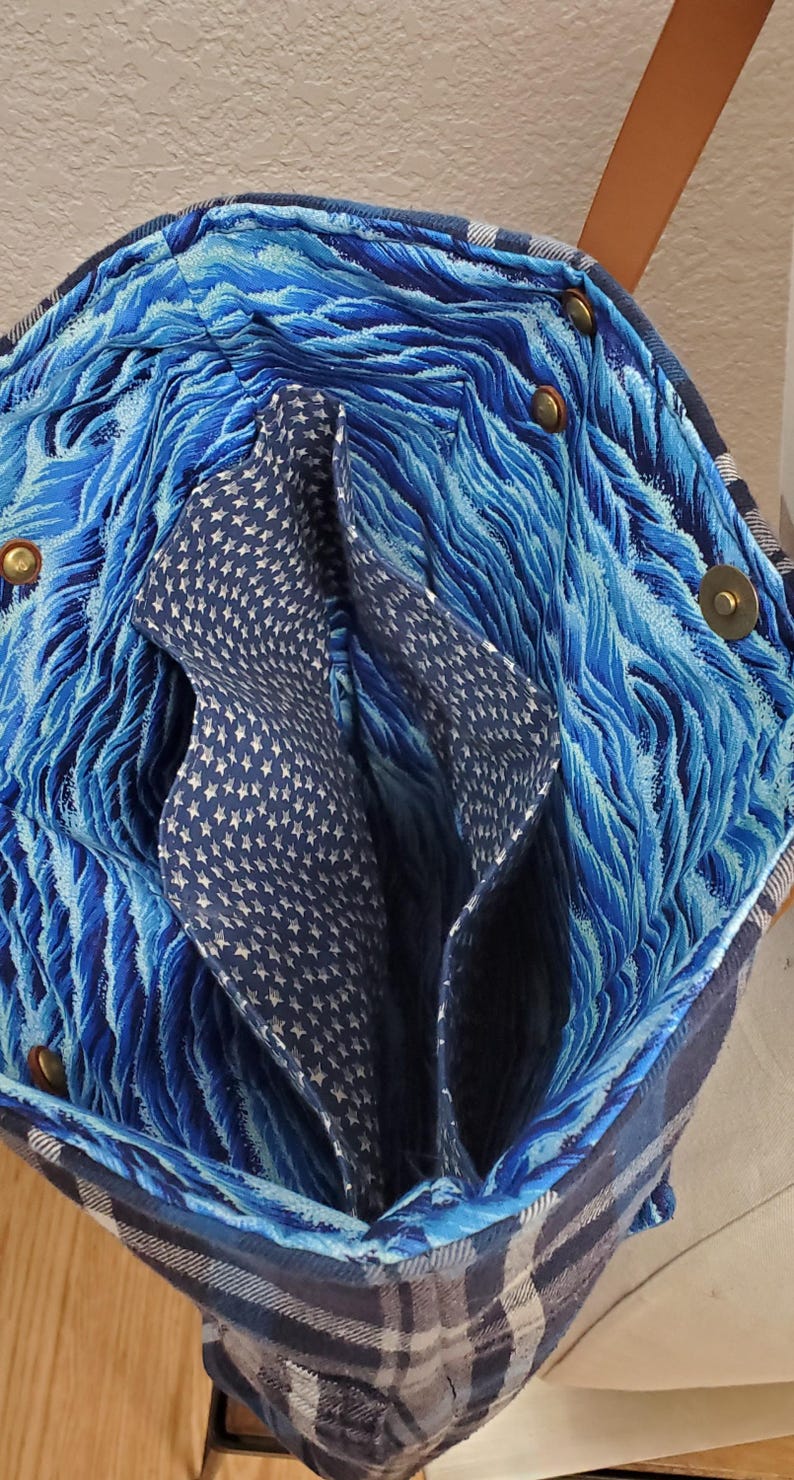 May include: A blue and white plaid tote bag with a blue and white wave pattern lining. The bag has a brown leather strap and brass rivets.