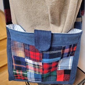 May include: A denim shoulder bag with a patchwork front featuring various plaid patterns in red, blue, and green. The bag has a blue denim flap and strap with a red and black plaid accent. The interior lining is white with blue stripes.