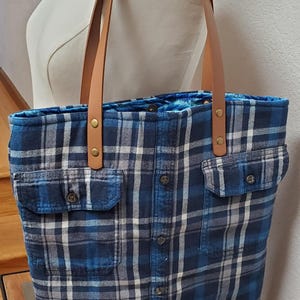 May include: A blue and white plaid tote bag with brown leather straps. The bag has two large pockets on the front and a button closure.