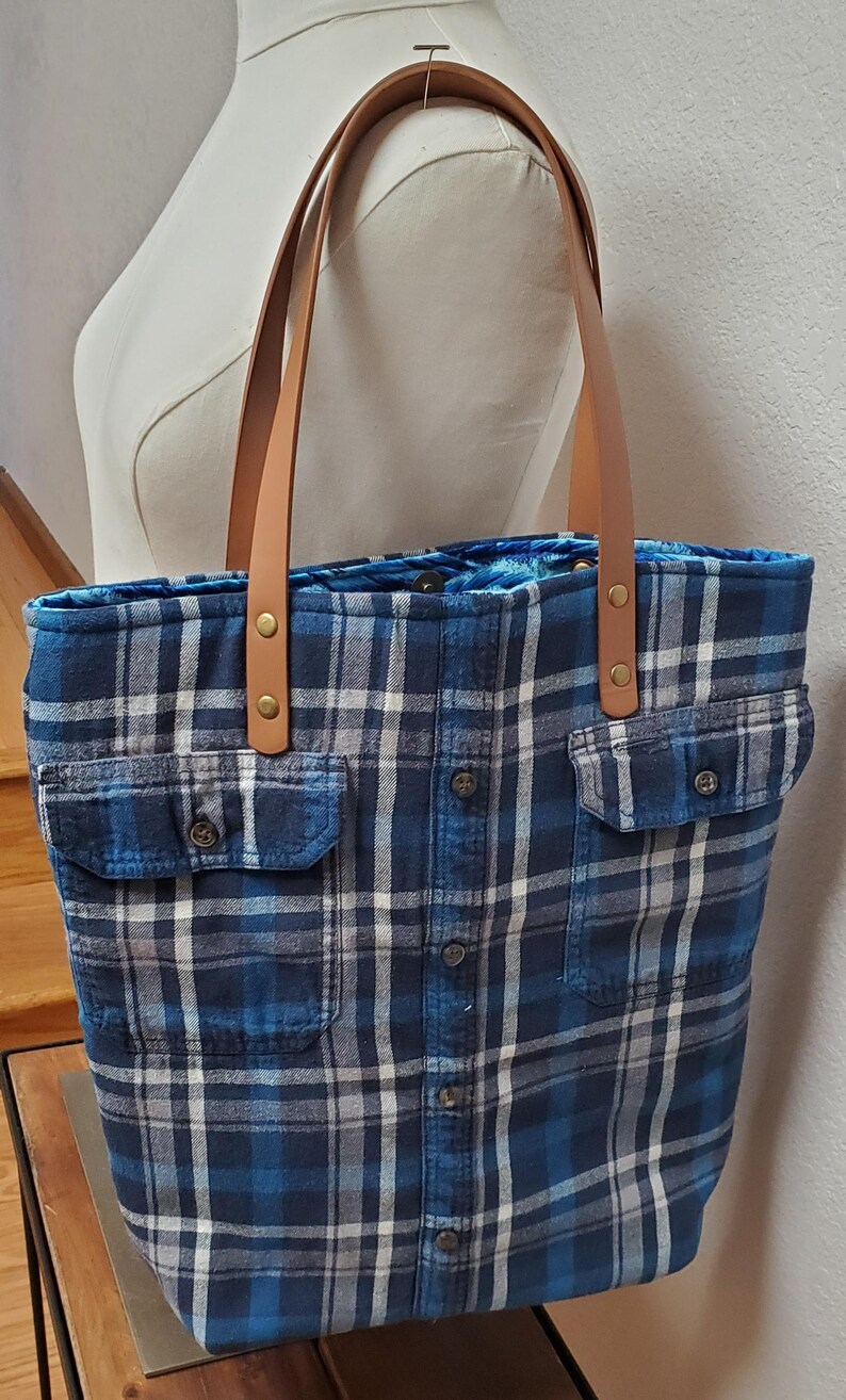 May include: A blue and white plaid tote bag with brown leather straps. The bag has two large pockets on the front and a button closure.