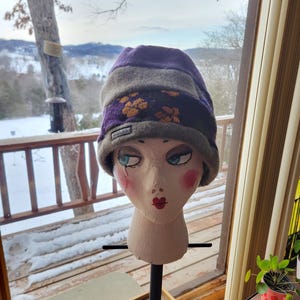 May include: A purple, gray, and brown knit beanie hat with a floral pattern. The hat is displayed on a mannequin head with painted features. The hat has the word "WALLES" on the brim. The background shows a snowy landscape.