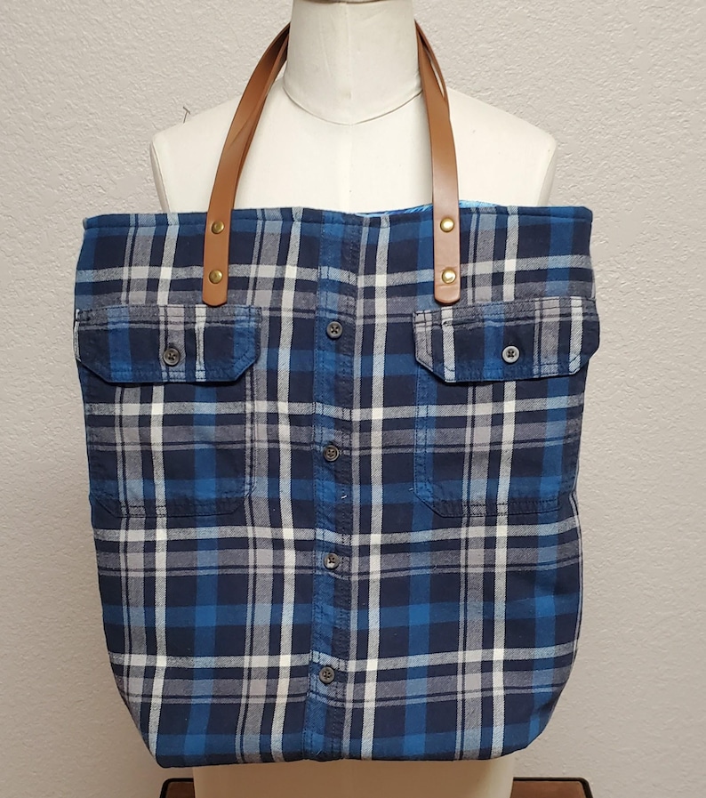 May include: A blue and white plaid tote bag with brown leather straps and gold-toned hardware. The bag has two large pockets on the front.