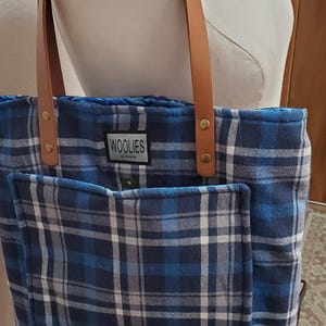 May include: Blue and white plaid tote bag with brown leather straps and a small pocket on the front. The bag has a label that says "Woolies" on it.