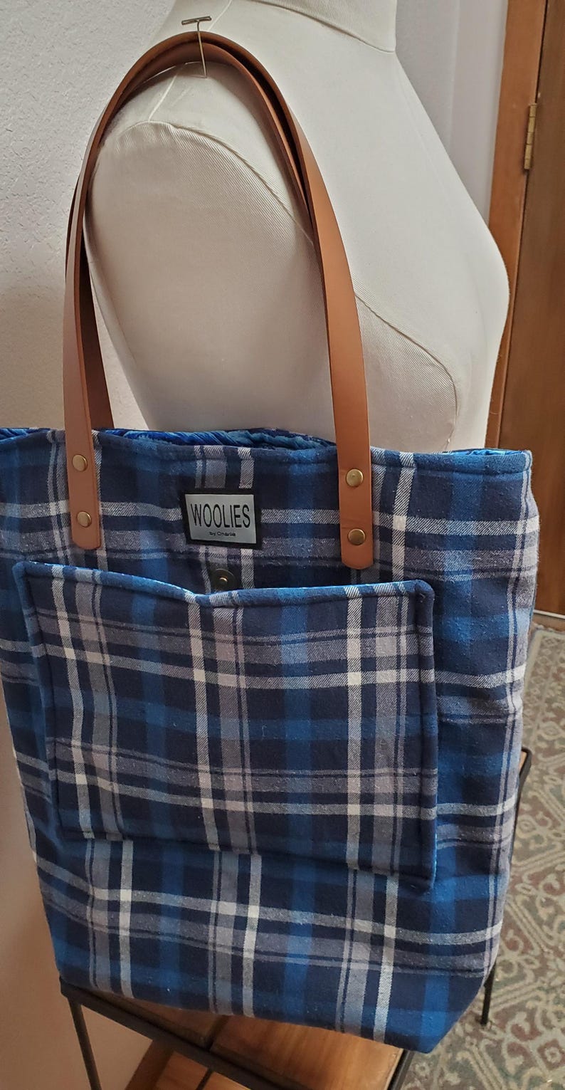 May include: Blue and white plaid tote bag with brown leather straps and a small pocket on the front. The bag has a label that says "Woolies" on it.
