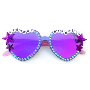 May include: Heart-shaped sunglasses with purple lenses and a light blue frame. The rims are decorated with rhinestones. The words "HIGHWAY" and "HYPNOSIS" are spelled out in white beads across the top of the lenses. Purple star accents are on the sides.