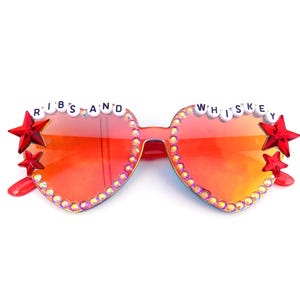 Può includere: Occhiali da sole a forma di cuore con lenti sfumate rosse. Le montature sono decorate con strass e stelle rosse. Il testo "RIBS AND WHISKEY" è scritto sulla parte superiore delle montature.