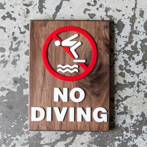 May include: A wooden sign with the words "NO DIVING" in white letters. A red circle with a diagonal line through it is above the text. The circle depicts a white silhouette of a person diving into water.