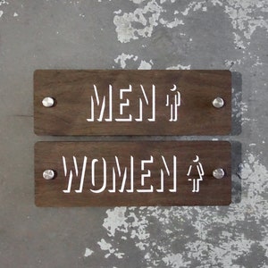 May include: Two wooden signs with the words "MEN" and "WOMEN" in white lettering. Each sign has a small silver screw on each side.