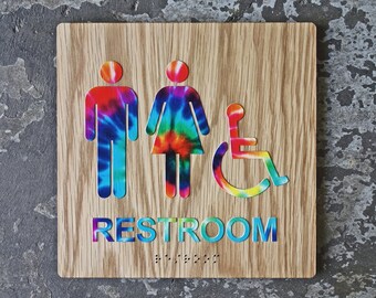May include: A wooden restroom sign with tie-dye symbols for men, women, and accessibility. The sign reads "RESTROOM" in blue and green letters.
