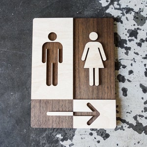 May include: A wooden sign with a white arrow pointing to the right. The sign is divided into four sections, with the top left section showing a male figure, the top right section showing a female figure, and the bottom sections showing the arrow.