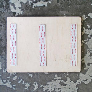 May include: A close-up of a light wood board with three strips of 3M VHB tape attached to the surface. The tape is white with red lettering that reads "3M VHB".