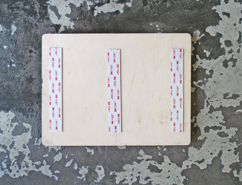 May include: A close-up of a light wood board with three strips of 3M VHB tape attached to the surface. The tape is white with red lettering that reads "3M VHB".