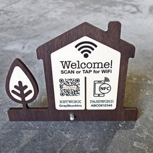 May include: Wooden house-shaped sign with a white background and black text. The sign says "Welcome! SCAN or TAP for WiFi". It also includes a QR code and a phone icon with the text "NFC". Below the QR code is the text "NETWORK GraySkunkInc" and below the phone icon is the text "PASSWORD ABCDE12345".