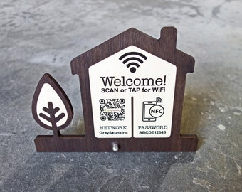May include: Wooden house-shaped sign with a white background and black text. The sign says "Welcome! SCAN or TAP for WiFi". It also includes a QR code and a phone icon with the text "NFC". Below the QR code is the text "NETWORK GraySkunkInc" and below the phone icon is the text "PASSWORD ABCDE12345".
