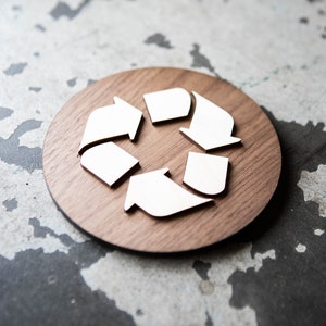 May include: A white recycling symbol made of wood, on a round wooden plaque. The plaque is brown and has a natural wood grain.