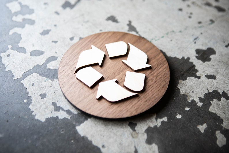 May include: A white recycling symbol made of wood, on a round wooden plaque. The plaque is brown and has a natural wood grain.