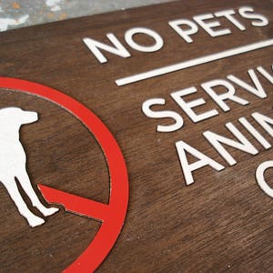 May include: A brown wooden sign with white text that reads "No Pets Service Animals Only". A red circle with a white silhouette of a dog is in the upper left corner of the sign.