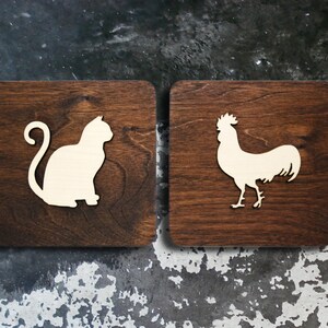 May include: Two square wooden signs with a dark brown stain. One sign features a white cat silhouette, and the other has a white rooster silhouette. The signs are likely for home decor or a rustic-themed setting.