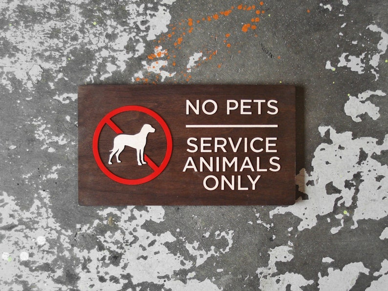 May include: A brown wooden sign with white text that reads "NO PETS SERVICE ANIMALS ONLY". A red circle with a diagonal line through it and a white silhouette of a dog is in the upper left corner of the sign.