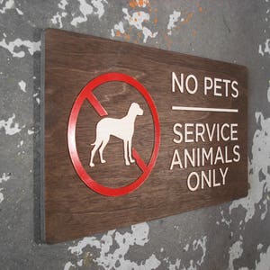 May include: A brown wooden sign with white text that reads "No Pets Service Animals Only". A red circle with a diagonal line through it and a white silhouette of a dog is in the upper left corner of the sign.