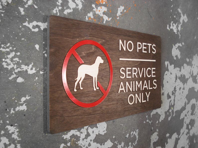 May include: A brown wooden sign with white text that reads "No Pets Service Animals Only". A red circle with a diagonal line through it and a white silhouette of a dog is in the upper left corner of the sign.