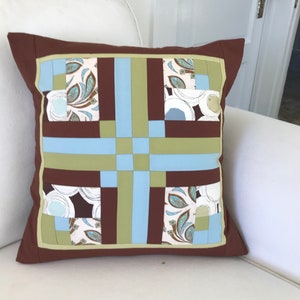 May include: A brown cushion with a patchwork design featuring squares of blue, green and white fabric. The cushion has a brown border.