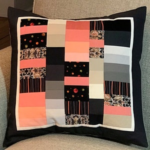 Peut inclure: Un oreiller noir avec une bordure blanche et un motif patchwork composé de carrés de tissu rose, gris et noir. Les carrés de tissu ont des motifs différents, notamment des pois, des rayures et des motifs floraux.