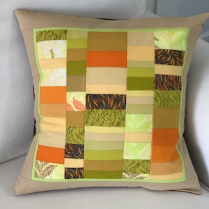 May include: A beige pillow with a brown border and a patchwork design of green, orange, and yellow fabric squares.
