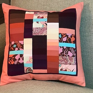 May include: A pink cushion with a patchwork design featuring various shades of brown, pink, and purple fabric. The cushion has a turquoise ribbon accent and a floral pattern.