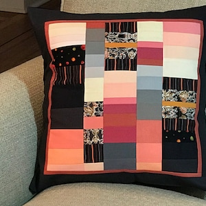 May include: A square cushion with a patchwork design featuring various shades of pink, red, grey, and black. The cushion has a black border with a red trim.