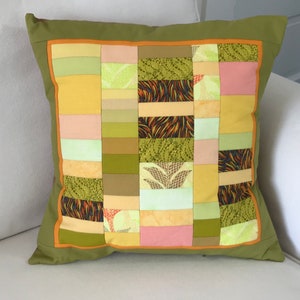 May include: A green cushion with a patchwork design featuring various shades of green, yellow, and pink fabrics. The cushion has a brown border.