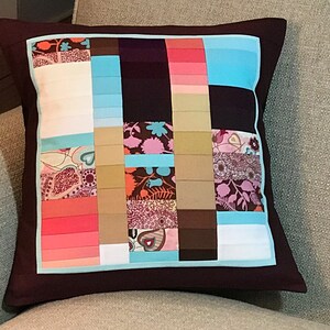 May include: A brown pillow with a blue border and a patchwork design featuring various shades of pink, brown, and beige. The patchwork design includes floral and geometric patterns.