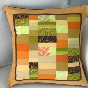 May include: A brown and tan patchwork cushion with a square pattern of different coloured fabrics. The cushion has a brown border and is made of a variety of fabrics, including green, orange, and tan.