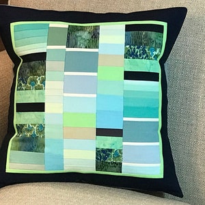 May include: Square decorative pillow with a navy blue border and a patchwork design. The patchwork features vertical stripes in shades of green, blue, and white, with some sections of patterned fabric. The pillow is resting on a neutral-colored sofa.