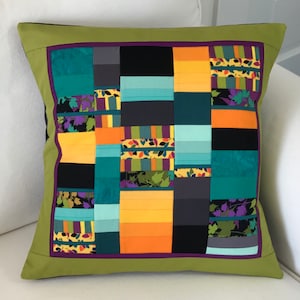 May include: A green cushion with a purple border and a patchwork design of black, orange, yellow, teal, and grey fabric. The fabric has a variety of patterns, including stripes, leaves, and geometric shapes.