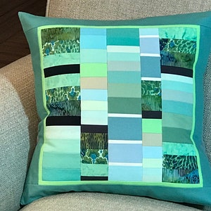 May include: A teal green cushion with a patchwork design featuring various shades of green, blue, and beige. The cushion has a light green border and a teal green backing.
