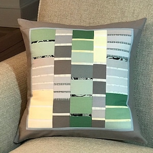 May include: A grey and white cushion with a patchwork design featuring squares of green, grey, and white fabric. The cushion has a light grey border.