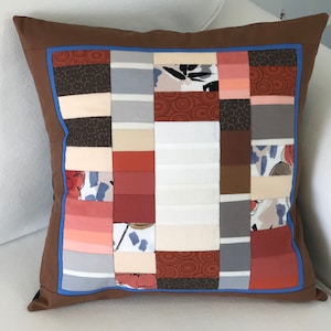 May include: A brown pillow with a blue trim and a patchwork design. The patchwork is made up of various shades of brown, gray, white, and pink fabrics with different patterns.