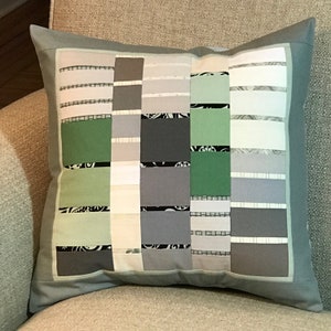 May include: A grey and green patchwork cushion with a geometric design. The cushion has a light grey border and is made of different fabrics with various patterns.