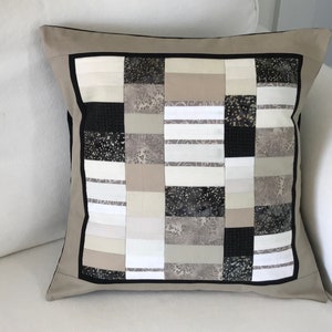 May include: A beige cushion with a black and white patchwork design. The cushion has a black border and is made of different fabrics.
